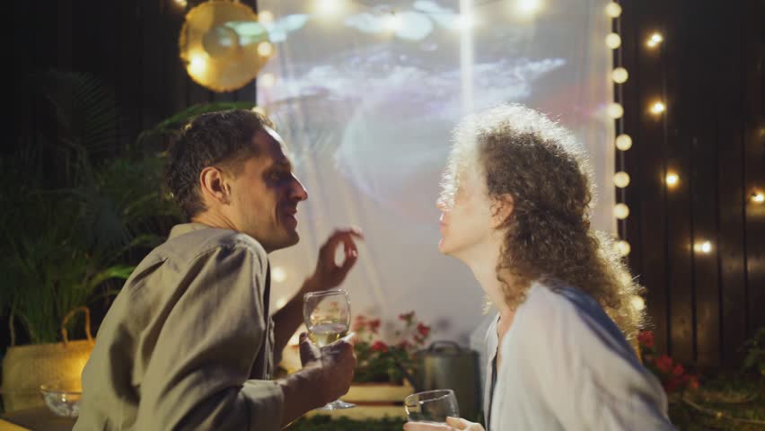 Close-up portrait man and woman with white wine glasses dancing opposite outdoor cinema screen, spending weekend at rest place with pillows and fairy lights in cottage yard Royalty-Free Stock Footage #3408815947