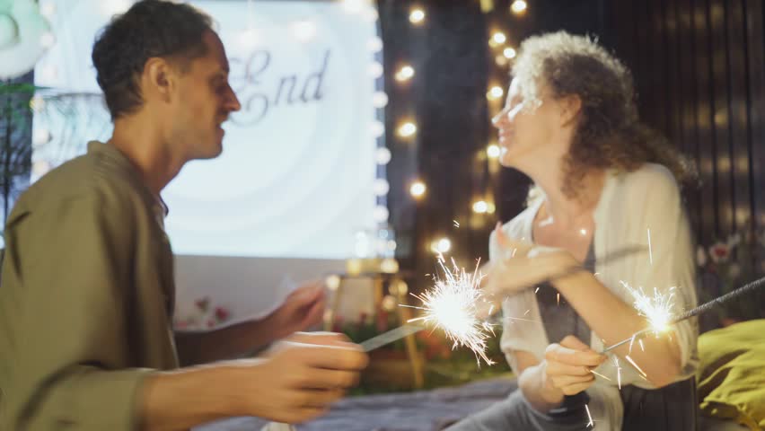 Close-up Lovely man and lady with sparklers in hands dance and have fun, celebrating and spending lazy weekend against bed sheet cinema screen , decorated fairy lights in the cottage yard Royalty-Free Stock Footage #3408818561