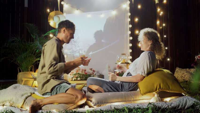 Lovely man and lady with white wine glasses dance and have fun, spending lazy weekend near large screen at rest place with pillows and fairy lights in cottage yard Royalty-Free Stock Footage #3408818859