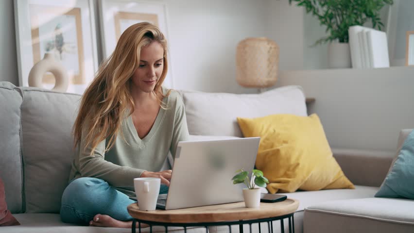 Video of beautiful woman working with her laptop while using mobile phone sitting on a couch at home.