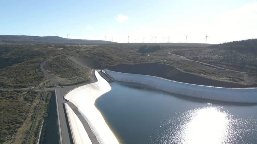 Aerial view of the Paul da Serra water reservoir built to harvest the rainwater. Drone rotating to the left showing the low level of water in it, the wind farm and the blue sky in the background.