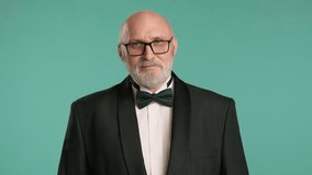Middle-aged man showing a gesture, lips sealed. He is dressed in a black tuxedo, white shirt with a bow tie. The man is bald with a gray beard, wearing glasses. Turquoise background.   8K RAW