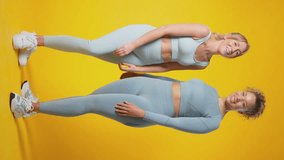 Vertical video full length studio portrait of two smiling women wearing gym fitness clothing exercising on yellow background - shot in slow motion