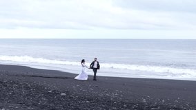 Drone footage of happy married couple walking, enjoying icelandic beauty of nature by the sea with rocks, huge mossy mountains, black sand with footprints. High quality 4k footage