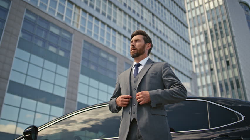 Millionaire posing at luxury car looking camera confidently zoom out. Elegant representative businessman standing at expensive auto near street skyscraper. Rich man buttoning jacket enjoying success.