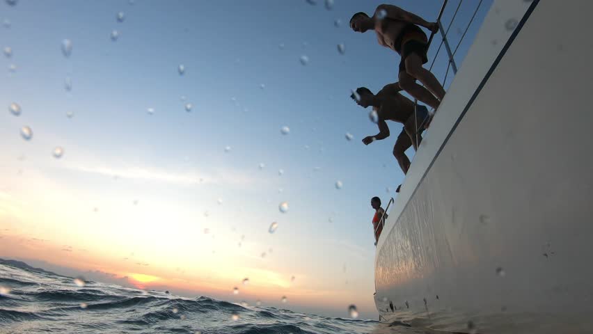 Group of young friends jumping to the beach, having a party in yacht. Attractive man and woman people hanging out, celebrating holiday vacation trip while catamaran boat sailing during summer sunset.