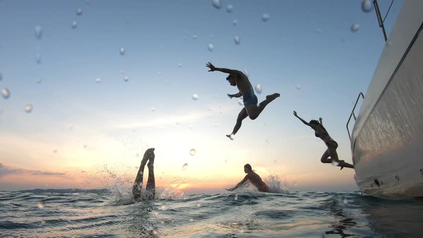 Group of young friends jumping to the beach, having a party in yacht. Attractive man and woman people hanging out, celebrating holiday vacation trip while catamaran boat sailing during summer sunset.