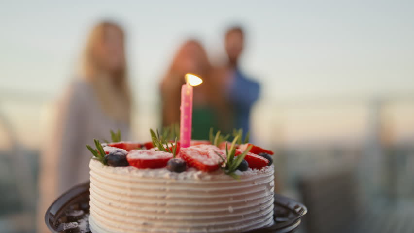 Friends bringing birthday cake to smiling woman at rooftop party close up. Excited girl blowing candle celebrating anniversary with mates. Laughing buddies making selfie happy surprise on smartphone.