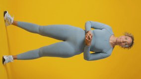 Vertical video studio full length shot of smiling woman wearing gym fitness clothing exercising on yellow background - shot in real time