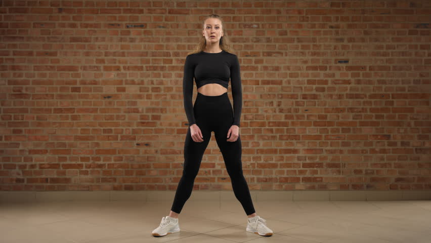 Woman coach engaged in sports against a brick wall background. She performs the exercise, sumo squat. The woman is focused and motivated to achieve good results. RED V-RAPTOR 8K RAW. Royalty-Free Stock Footage #3409175393