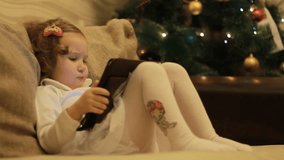 A little cute girl lies in sofa in a living room near Christmas tree, looking cartoon and playing the game. Funny child looks at the screen and plays downloaded application on tablet, laptop, computer