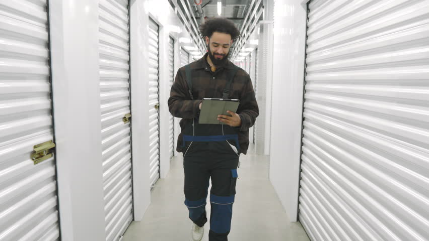 Young male worker of climate controlled storage unit examinating modern specialized facilities walking along long white secured corridor using gadget. High quality 4k footage