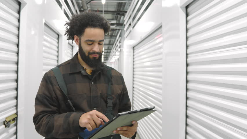 Happy dark-skinned man working in climate controlled storage units, clicking on tablet looking at shutter door, nodding approvingly, well lit interior. High quality 4k footage Royalty-Free Stock Footage #3409238767