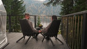 Husband pets wife hand sitting in armchairs. Woman talks to man on hotel terrace holding hands against river and mountains. Eco-friendly glamping resort - Powered by Shutterstock - Get 15% off with code: PIKWIZARD15
