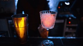 Barman pouring orange red alcohol cocktail with ice on a bar with neon lights at night