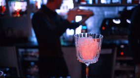 Barman shaking and pouring orange red alcohol cocktail with ice on a bar with neon lights at night