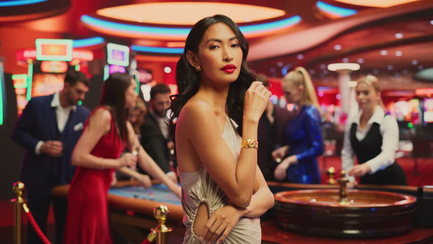 Portrait of a Beautiful Young Asian Woman, Wearing an Elegant Silver Dress, Posing in a Glamorous Atmosphere of a Casino, Surrounded by Active Gamblers Playing Roulette On the Background. Slow Motion