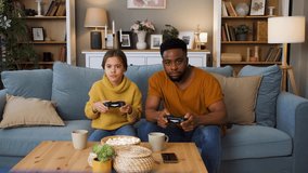 Father and daughter playing video game at home white sitting on sofa in living room.
