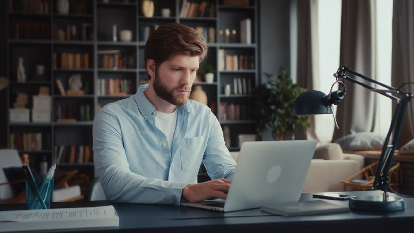 young man with beard and in blue shirt sits at table in home office and works at laptop, looks attentively with tension at screen, reads bad news, closes laptop, stands up and goes away quickly Royalty-Free Stock Footage #3409562549
