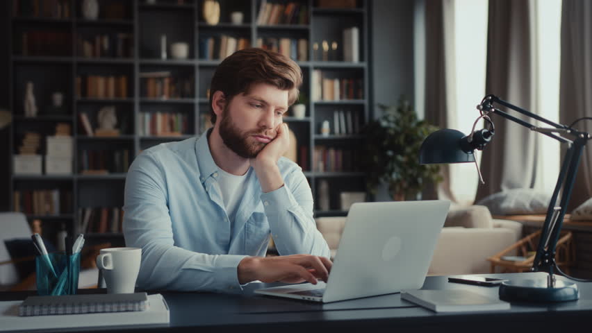 young tired and burn out man in blue shirt sits at table in home office and works at laptop, bored and unmotivated types slowly on keyboard without energy and interest, procrastinating Royalty-Free Stock Footage #3409574873