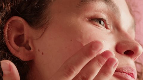 Young woman applying hydrogelic serum cream on her face close-up. Morning skin care routine. Pink background Adlı Stok Video