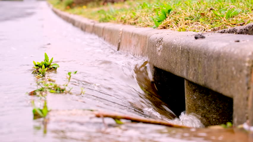 Rainwater runoff torrent runs down concrete gutter into stormwater road drain