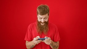 Confident young redhead man smiling happily as he plays his favorite video game, standing casually over a bold, isolated red background, fully immersed in the thrill of his mobile entertainment