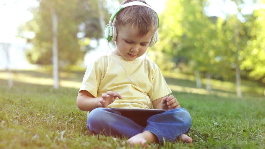 Cute little boy with headphones and a Tablet PC sitting on the grass. Online.
