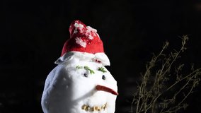 Snowman in the night. Home made snow man in the backyard garden with red scarf, Christmas hat, carrot nose, rock teeth. Beautiful winter holidays scene. 4k video tilt camera movement.