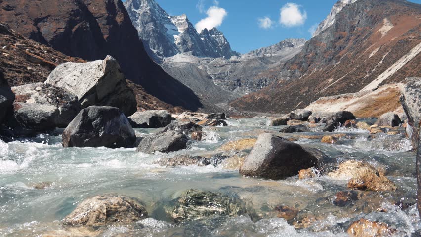 Clear stream running over stone boulders in highlands of Himalayas. River flowing along stone bottom against background rocky snowy mountains. Water of mountain river splashes on sunny summer day Royalty-Free Stock Footage #3410178429
