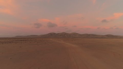AERIAL: Flying towards a large volcanic structure in pink colored desert sunset. Barely visible dirt tracks running through empty landscape. Quiet and colorful summer sunset in lonely volcanic desert.