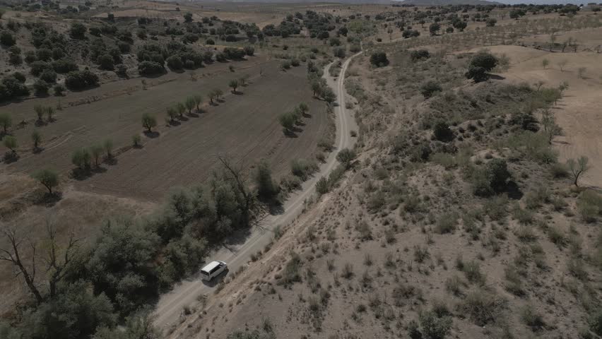 aerial view of a digital nomad driving on a narrow empty road between deserted hills covered with dry grass and trees under sunlight