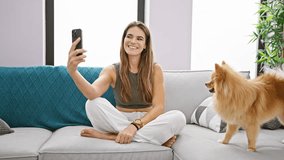 Relaxed young hispanic woman enjoying a funny video call on her mobile with her happy dog by her side, sitting on the living room sofa at home.