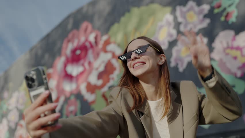 Happy smiling woman talking on smartphone by video call, waving hand on camera while walking in city outdoors. Communication online concept. Stylish girl blogger in trendy pixel glasses with phone