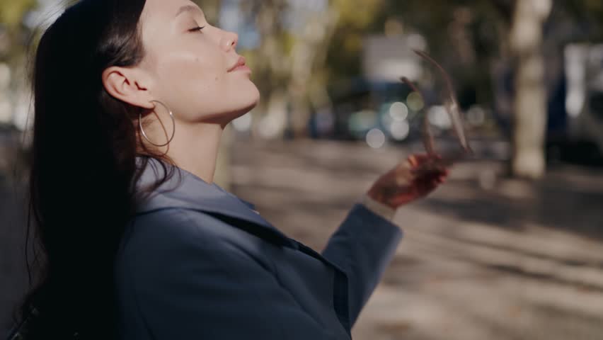 Attractive woman put on sunglasses and looking at camera in street. Portrait of stylish female in trendy glasses outdoors in sunny day.