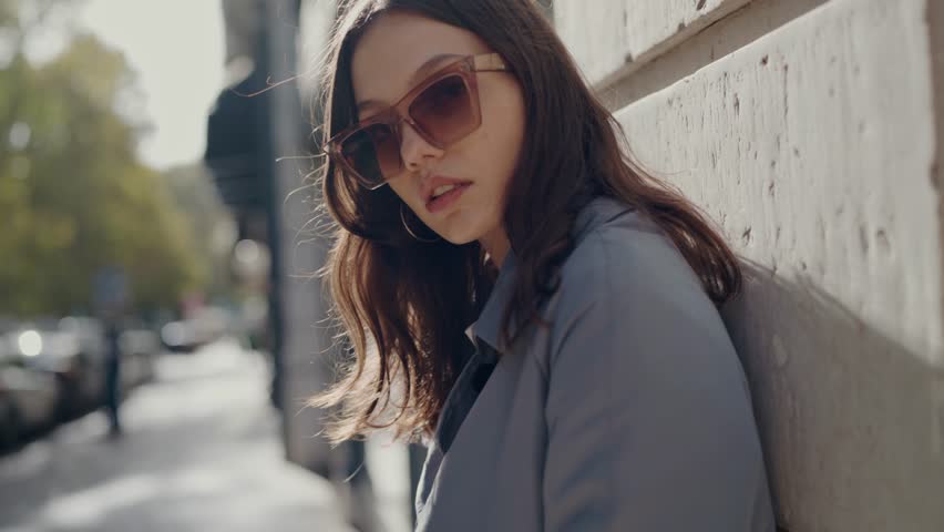 Young attractive woman in trendy sunglasses posing on camera while lean on building wall at city outdoors. Portrait of female in stylish outfit on street