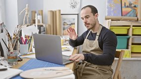 A bearded man in a bib apron smiles during a video call in a creative art studio, surrounded by paintings and art supplies.