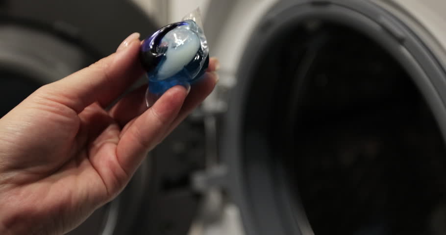 Close up woman puts colorful gel capsules with laundry detergent in the washing machine on dirty laundry. Modern liquid detergents.