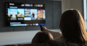 Woman selects tv channels with remote control at living room on sofa. Woman with her little daughter controls TV using a modern remote control.