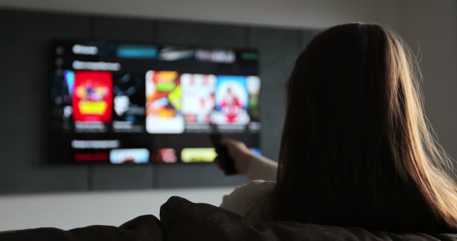 Woman selects tv channels with remote control at living room on sofa. Woman controls TV using a modern remote control.