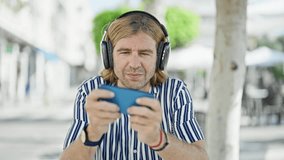 A middle-aged man with headphones uses a smartphone on a sunny urban sidewalk, expressing focus and technology engagement.