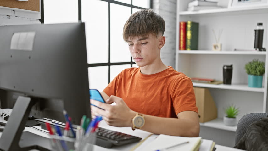 Young caucasian male student in orange shirt takes selfies at a modern university library desk with smartphone.