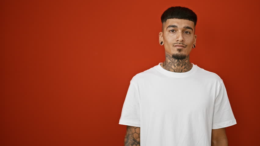 Joyful young latin man confidently presents with pointing fingers over a vibrant isolated red background, revealing his tattooed hands while showcasing a self-assured, infectiously happy smile.