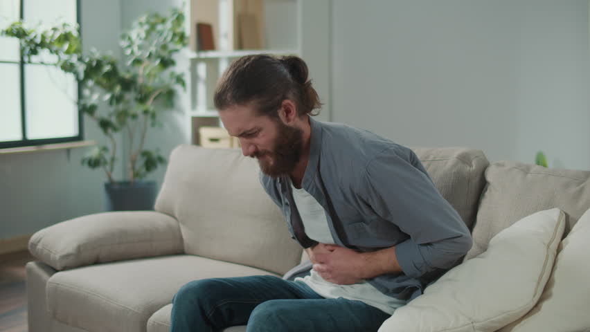 Man sitting on the couch suddenly felt pain in his stomach. Man trying to relieve pain by massaging his stomach Royalty-Free Stock Footage #3410498325