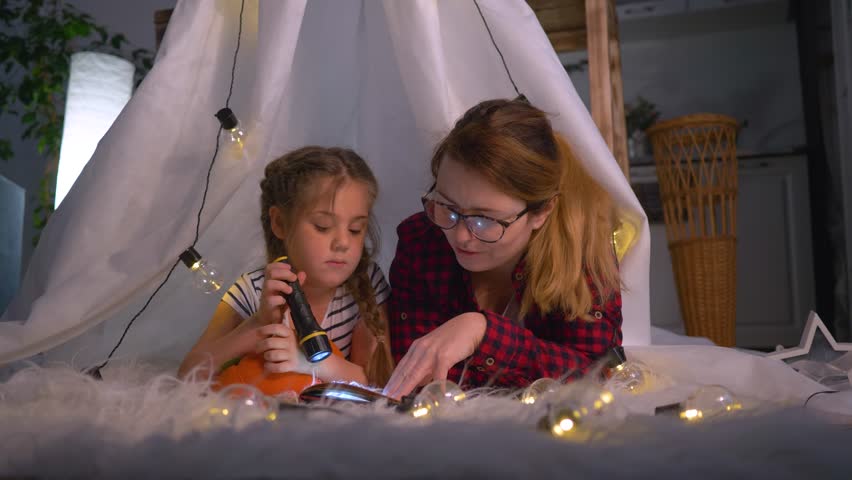 A happy family. Mom and daughter are reading a book in the children's room tent. Mother and daughter with a book and a flashlight before bed at home. Happy family lifestyle Royalty-Free Stock Footage #3410585785