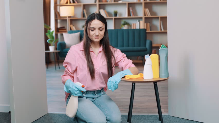 a girl in a pink shirt wearing big rubber gloves sits on the floor at home and poses for the camera. Female with detergents at home in the room