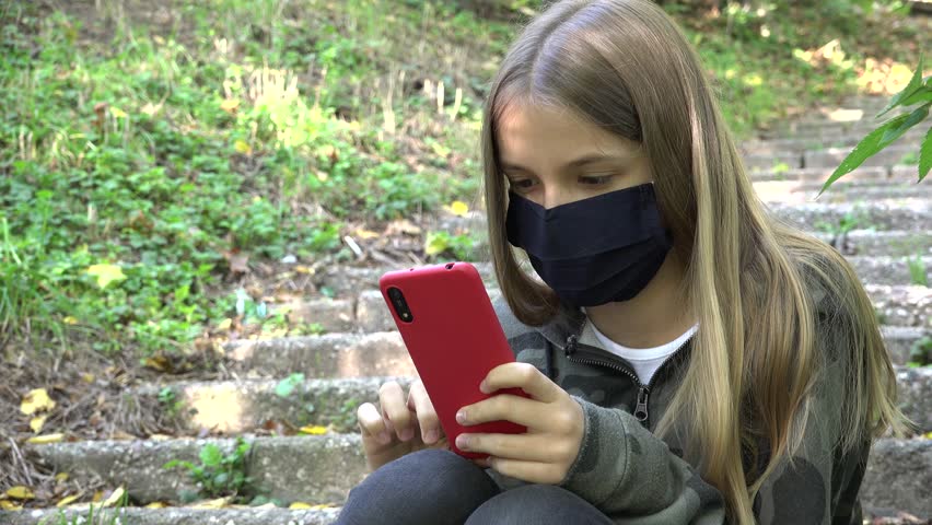 Sick Child with Protective Mask due Coronavirus Pandemic, Kid Browsing Internet on Smartphone in Park, Girl Playing Smart Phone