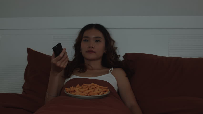 Medium shot of gen Z girl of Asian ethnicity lying in bed while watching TV shows and eating chips or snacks at nighttime, enjoying spending time alone
