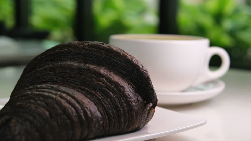 black croissant with cuttlefish ink on a white plate with a cup of cappuccino latte coffee against a green window background summer breakfast, France gourmet food delicious lunch snack