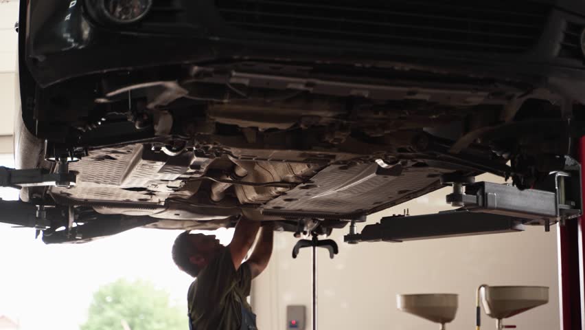 Auto mechanic works on car suspension in repair shop. Fixes vehicle underside, inspects springs, shock absorbers. Professional garage service, maintenance. Car lifted, technician examines chassis.